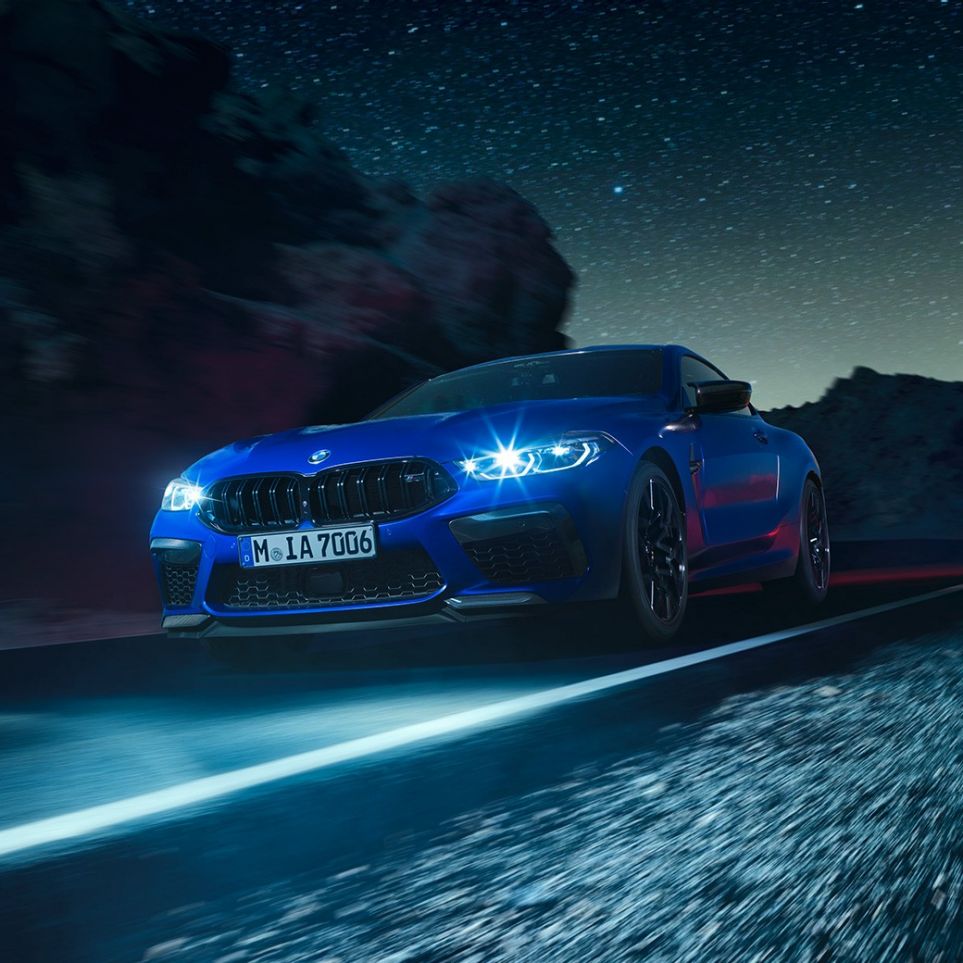 Driving BMW M8 Coupé, three-quarter front view. BMW M8 Coupé in Marina Bay Blue metallic with illuminated LED headlights, three-quarter front view, driving at night.