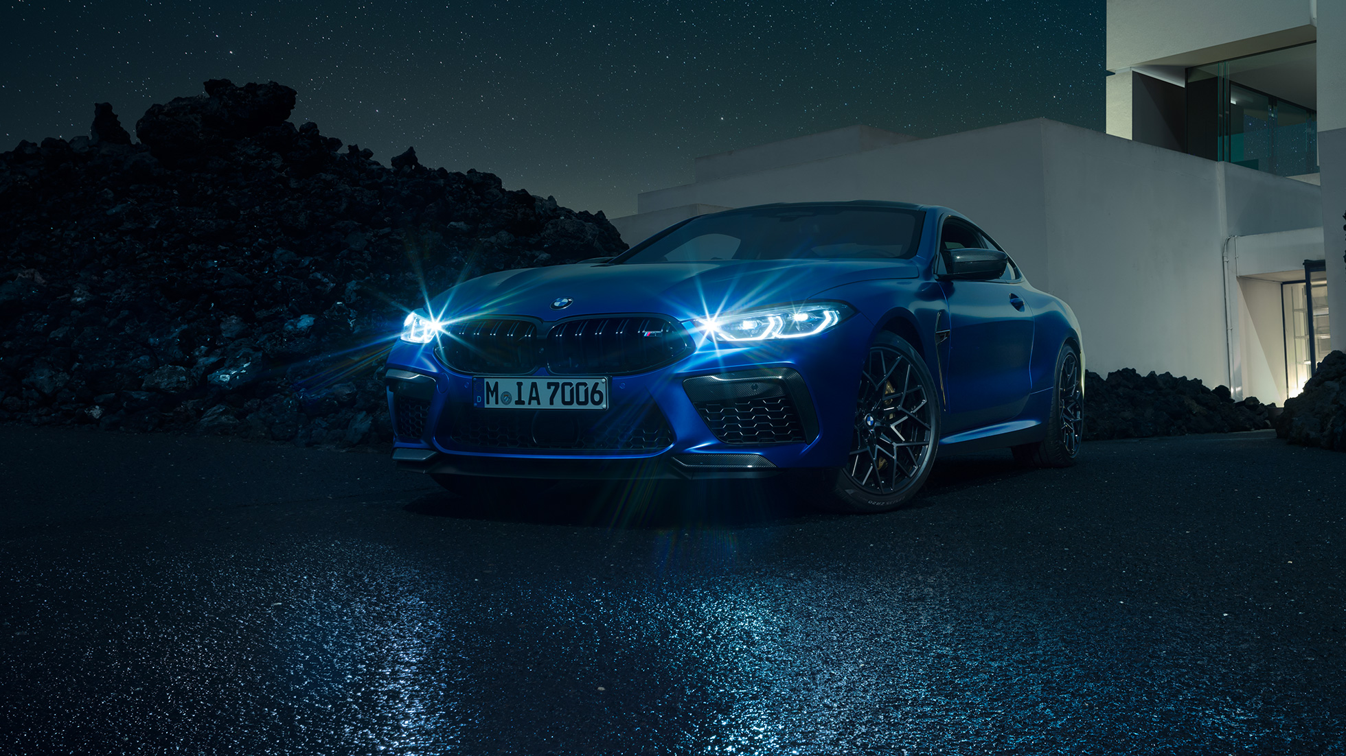 BMW M8 Coupé, three-quarter front view. BMW M8 Coupé in Marina Bay Blue metallic with shining LED headlights, standing three-quarter front view in front of villa at night.