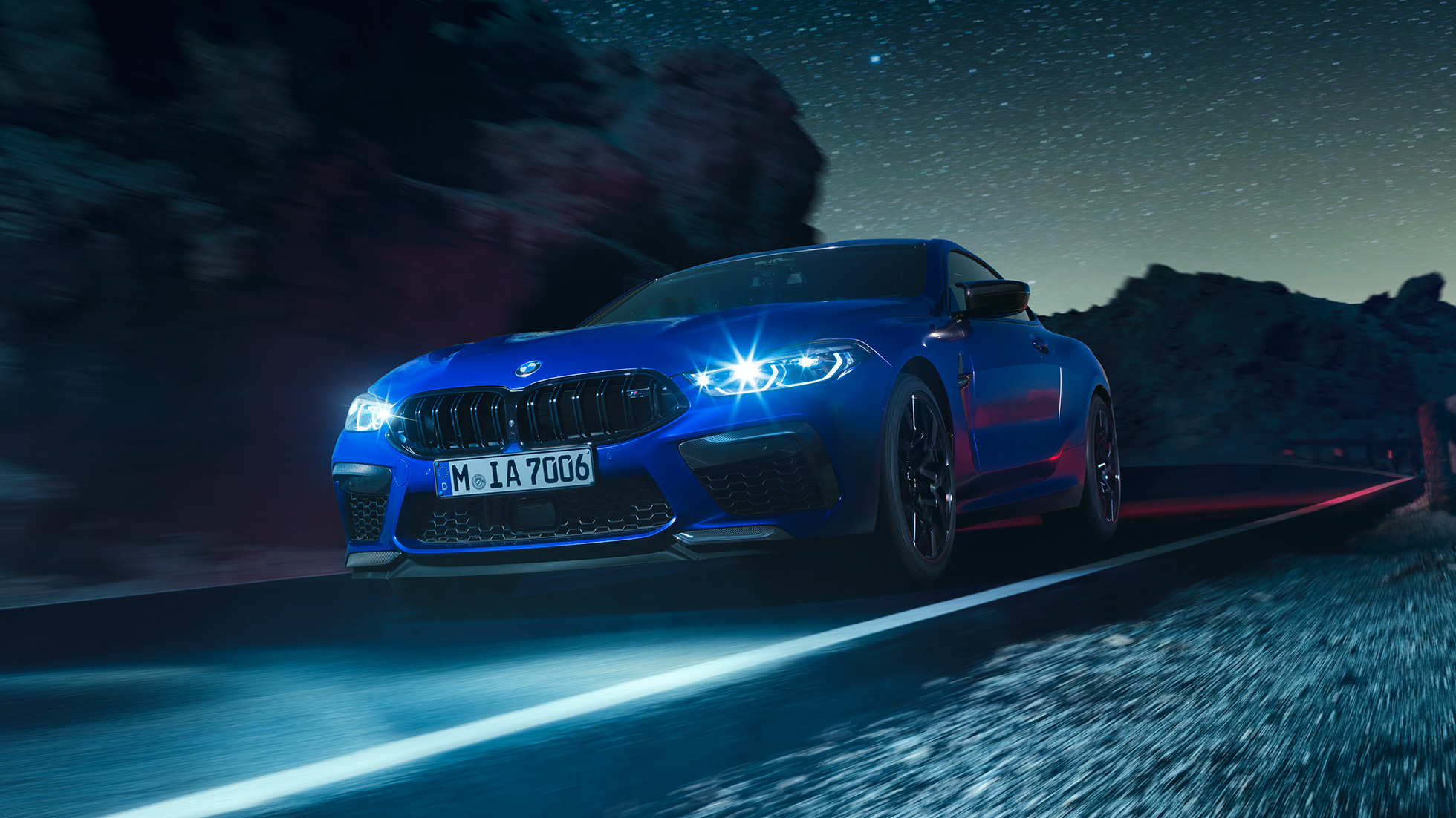 Driving BMW M8 Coupé, three-quarter front view. BMW M8 Coupé in Marina Bay Blue metallic with illuminated LED headlights, three-quarter front view, driving at night.