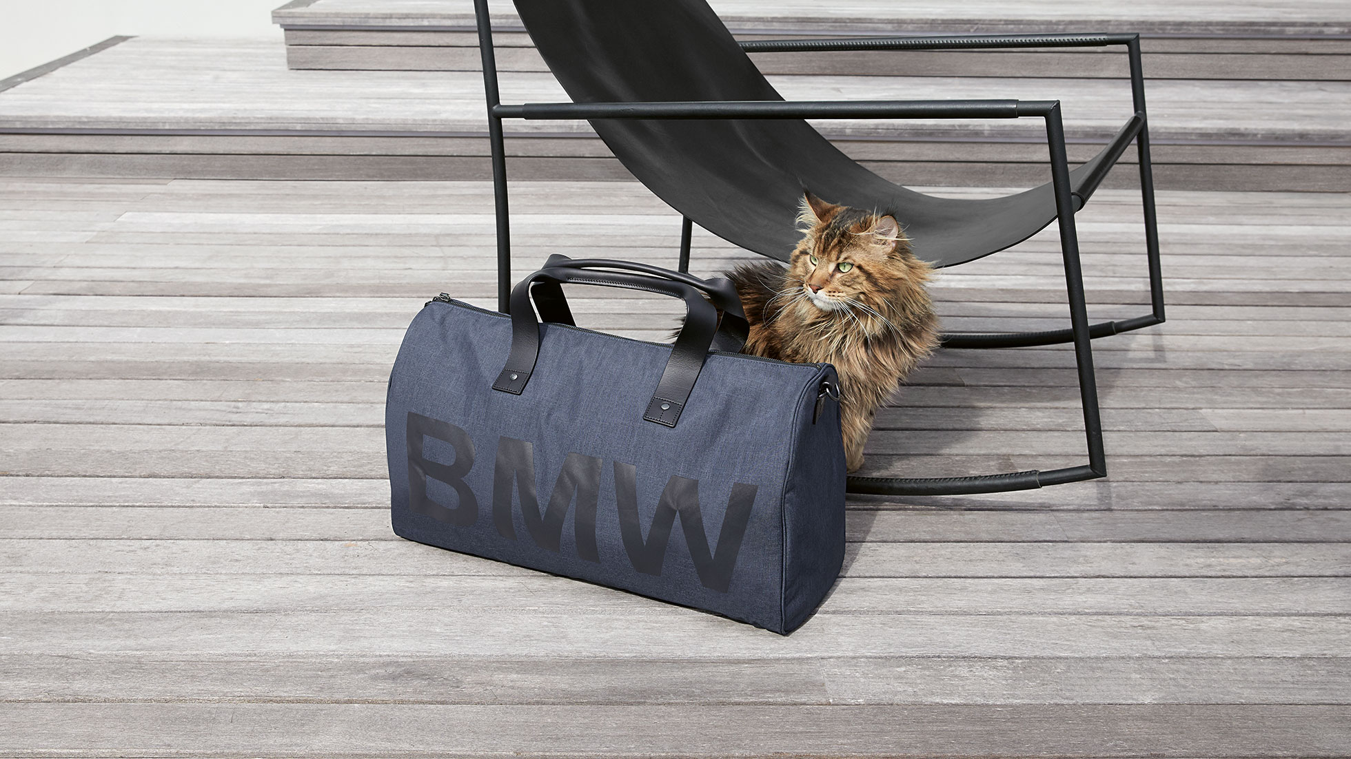 The picture shows the BMW Modern Duffle Bag in front of a sitting cat and a chair. The picture shows the BMW Modern Duffle Bag in front of a sitting cat and a chair.