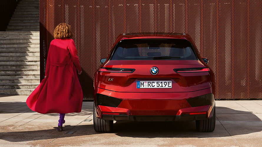 Closing the tailgate Woman in red coat next to BMW iX (i20) in front of building